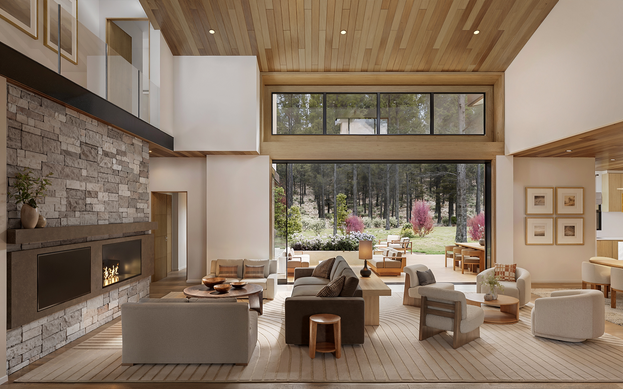 3D interior rendering of the Pine Cone Road double-height living room, with a floor-to-ceiling stone fireplace and a linear gas firebox, a wood-slat vaulted ceiling, a grouped seating arrangement around a round wood coffee table, and a wall of glass opening to the pine-edged backyard