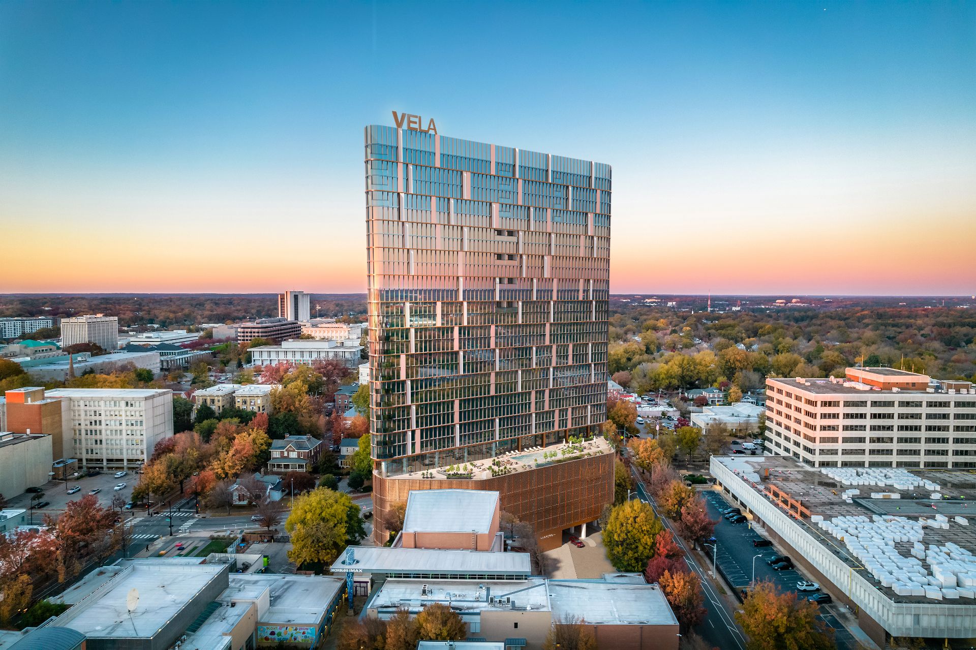 Aerial 3D rendering of the VeLa mixed-use tower in Raleigh at dusk, its gridded glass facade lit with rooftop VeLa signage above the downtown skyline and autumn tree canopy.